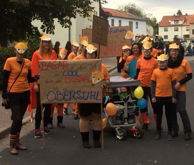 Foto vom Kirmes-Umzug: Menschen mit orangenem T-Shirts, Fuchs-Maske und Schildern (vorne: Basar in jedem Ort, wie cool, doch das Original ist in Obersuhl)