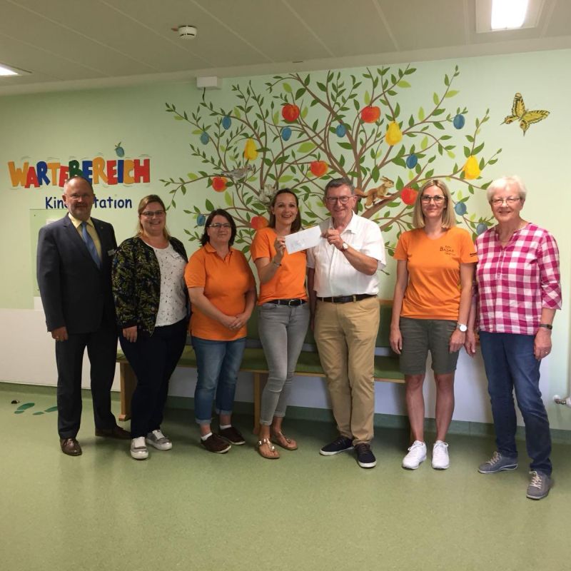 Spenden-Übergabe: sieben Menschen vor einer Wand stehend (Wartebereich Kinderstation)